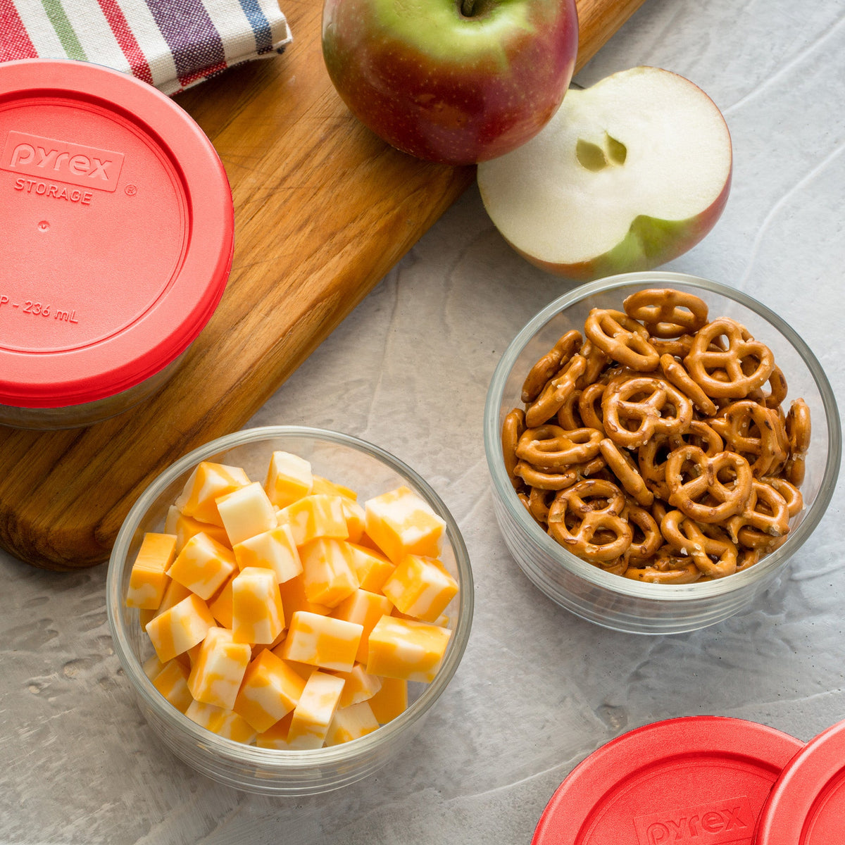 Bowl of cheese cubes, pretzels, and half an apple on a gray surface with Pyrex containers.